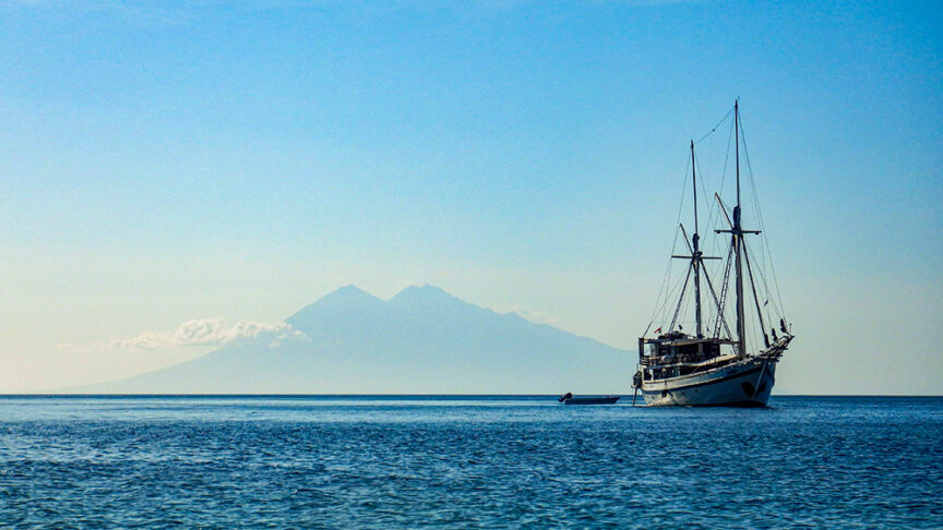 A sailboat is anchored in calm blue waters with a distant mountainous island of Komodo visible under a clear sky, setting the scene for an unforgettable multi-adventure trip.