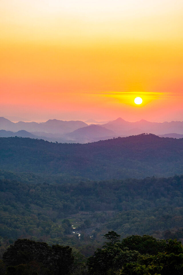 A bright sun sets over a range of mountains covered with dense forest, casting an orange and yellow glow across the sky, reminiscent of our last multi-adventure trip to Komodo.