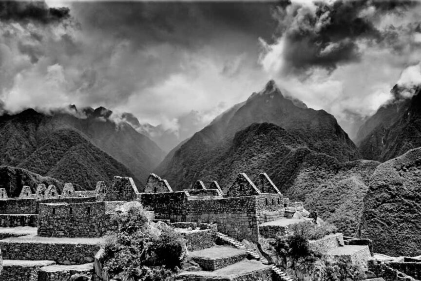 Black and white image of Machu Picchu ruins set against a dramatic, cloudy mountain backdrop. The ancient stone structures are terraced on a hillside, framed by mist-covered peaks. This picture is truly worth a thousand words.