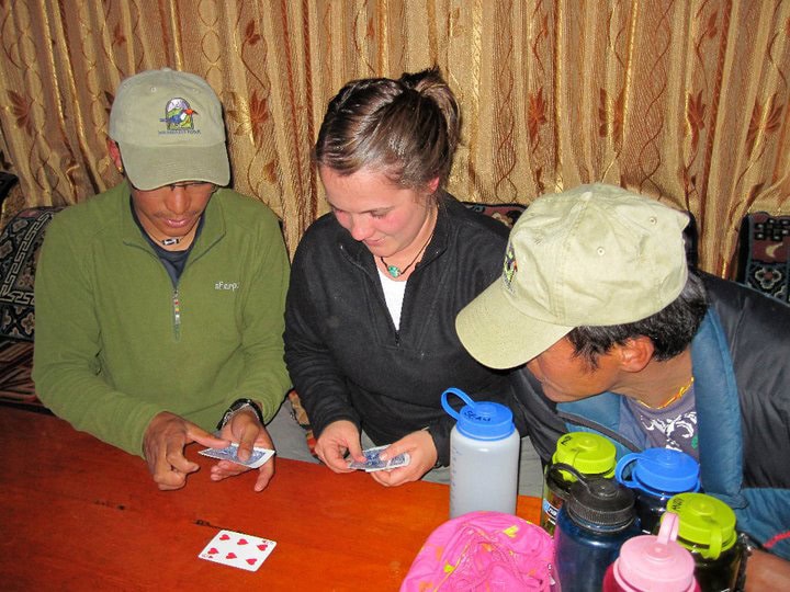 Three people in outdoor attire and caps sit at a table playing cards, surrounded by water bottles and gear. Their laughter echoes like a moment captured from a Nepal adventure photobook, blending the warmth of family with the thrill of exploration.