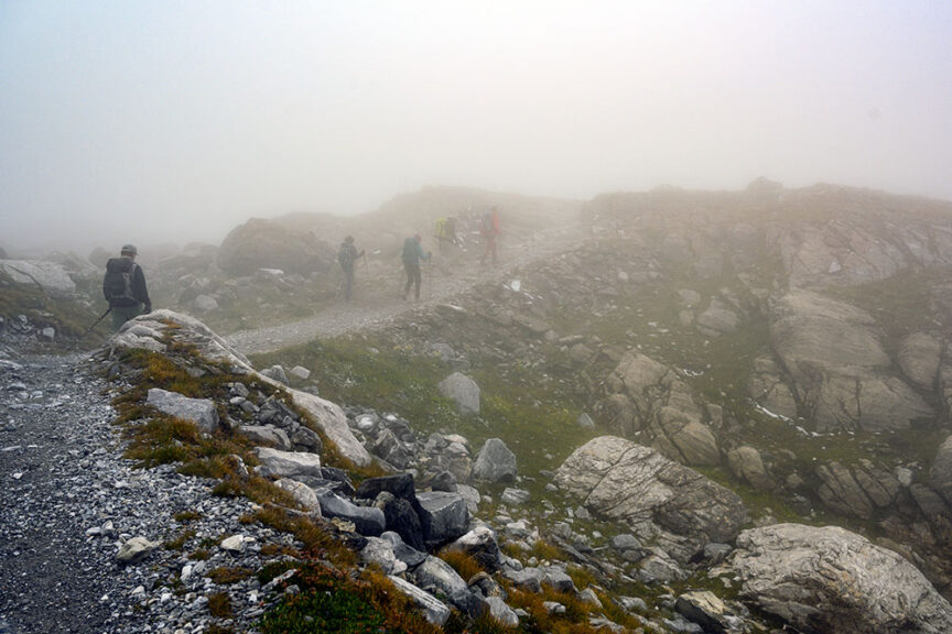 Several hikers walk along a rocky mountain trail in dense fog, surrounded by boulders and sparse vegetation, experiencing the challenging terrain of the GR5 route, often referred to as the journey from Mountains to the Sea.
