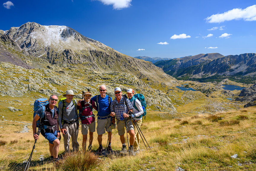 A group of hikers stands on a grassy slope along the GR5 trail, with majestic mountains and a sprawling valley in the background under a bright blue sky.