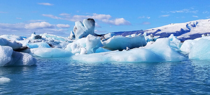 Numerous icebergs float in a calm, bright blue glacial lake under a clear sky with scattered clouds, perfect for hikes. Snow-covered mountains are visible in the background, reflecting Iceland's pristine beauty.