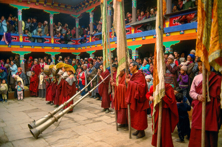 A group of people in red robes play long horns in a courtyard filled with onlookers during an amazing cultural event at a brightly decorated, multi-story building.