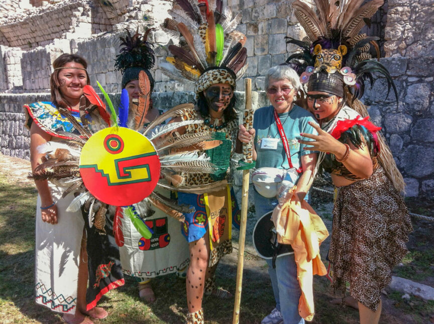 A group of people wearing traditional indigenous attire, featuring feathers and bright colors, pose alongside another person wearing casual clothing in front of ancient stone ruins. The scene evokes the essence of the Maya civilization, as if unlocking secrets from a bygone era.