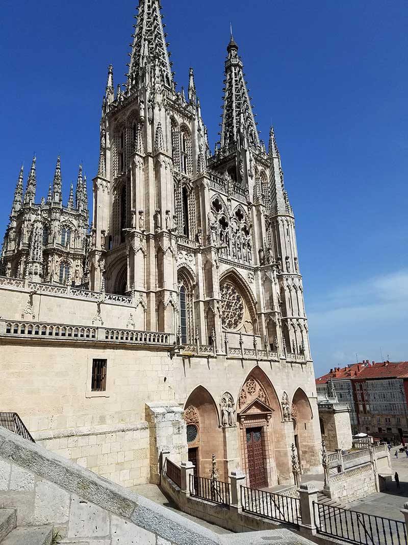 A large Gothic-style cathedral with intricately detailed spires and arches under a clear blue sky, reminiscent of the magnificent stops along the Way of St. James in Spain. Steps lead up to the main entrance, inviting pilgrims and travelers alike to explore its grandeur.