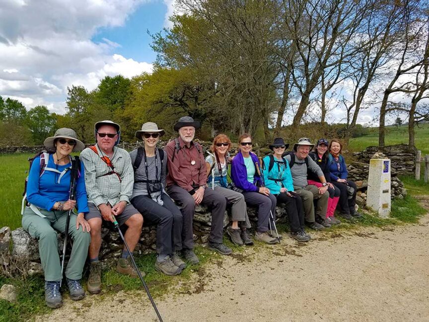 A group of nine people, dressed in outdoor gear, are seated on a stone wall in a rural area with trees and grass in the background. They are smiling and appear to be on a pilgrimage, possibly along the Way of St. James in Spain.