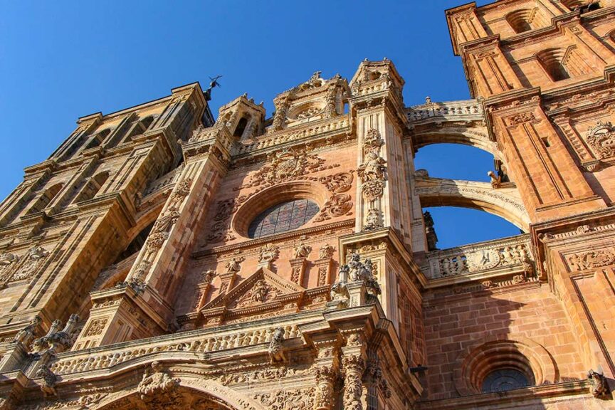A close-up view of the ornate facade and towers of an old stone cathedral with detailed carvings and sculptures against a clear blue sky, marking a significant stop on the Way of St. James pilgrimage in Spain.