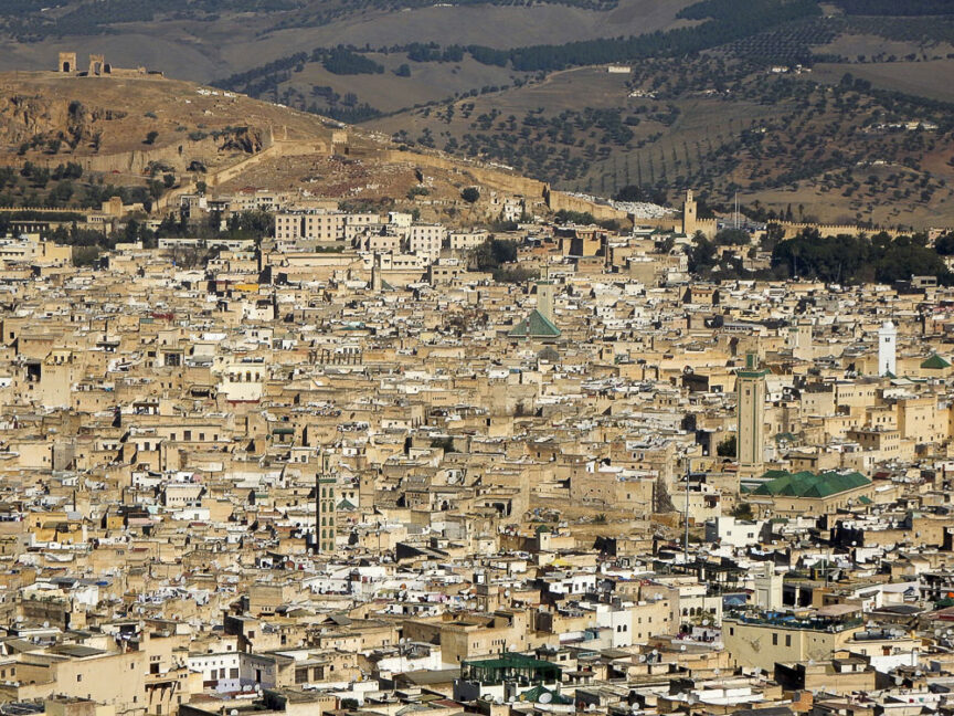 Aerial view of a densely built beige city in Morocco, featuring various buildings, minarets, and surrounding hills.