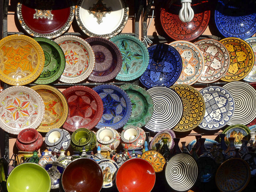 A collection of colorful, intricately patterned ceramic plates, bowls, and cups reminiscent of a Moroccan market displayed on shelves.