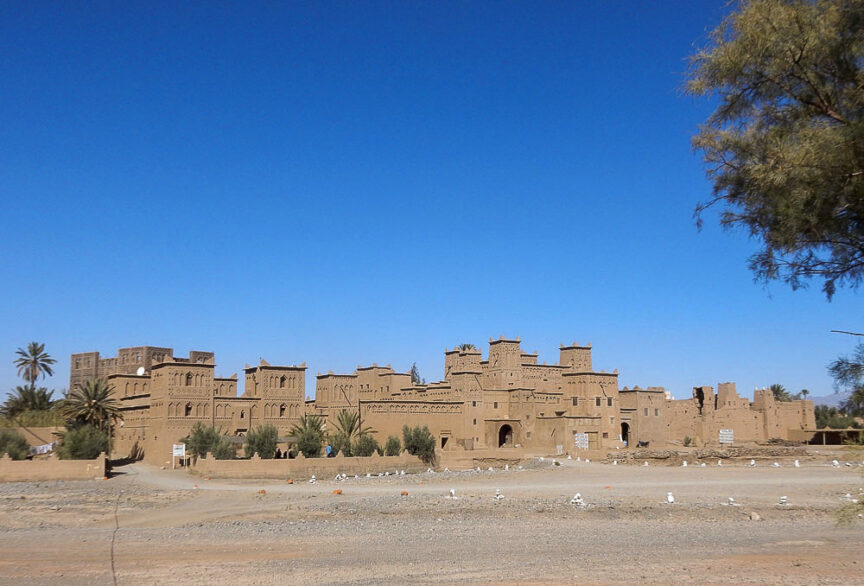 A large, traditional earthen structure with multiple towers and walls stands under a clear blue sky along the coast of Morocco, with some vegetation and open land in the foreground.
