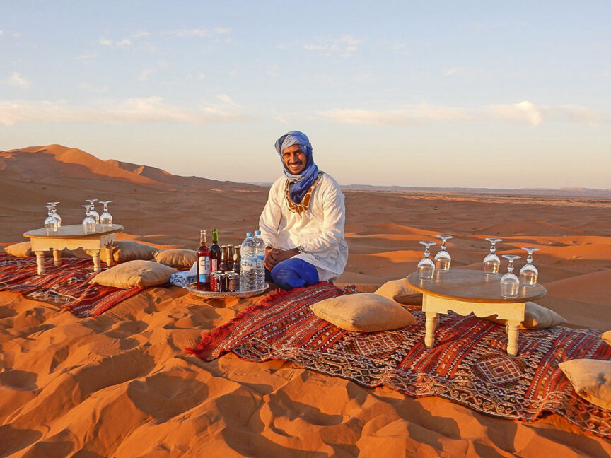 A traveler in traditional Moroccan attire sits on a rug in the desert with drinks and glassware arranged on small tables. Sand dunes stretch into the distance.