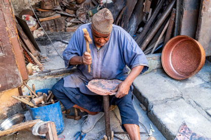 A man sits on the ground in a workshop while hammering a metal piece, reminiscent of an adventurer forging his own path. Various tools and materials are scattered around him, evoking the rugged spirit of hiking through Morocco's vibrant landscapes.