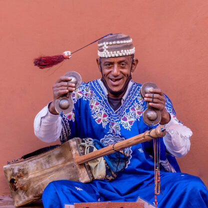 A person wearing traditional clothing and a decorated hat is seated against a red wall in Morocco, smiling and holding clapping cymbals. They have a stringed instrument resting on their lap, reminiscent of tales from an adventurous journey through Moroccan landscapes.