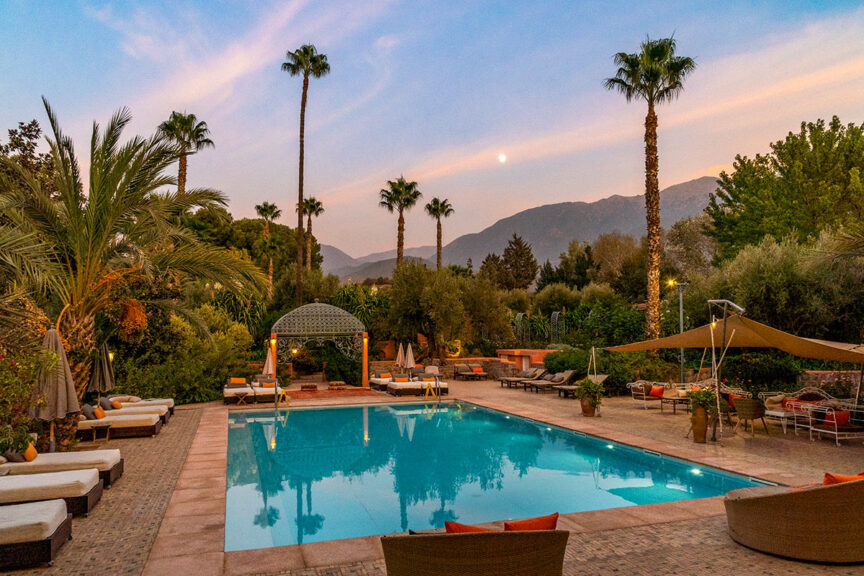 A serene outdoor pool area with lounge chairs, palm trees, and mountains in the background at sunset invites you to unwind after a day of adventure hiking in Morocco.