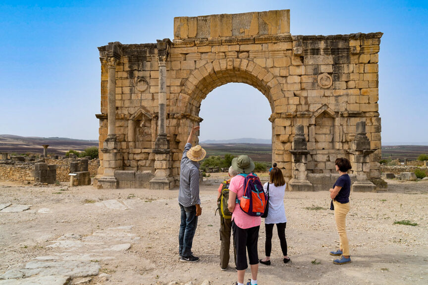A group of four people stand near a large ancient stone archway in Morocco, with one person pointing towards it. The background features a dry, open landscape under a clear blue sky, capturing the spirit of adventure and hiking through this breathtaking scenery.