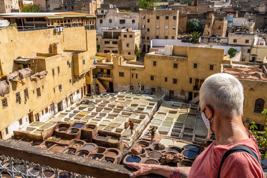 A person with short gray hair, wearing a pink shirt and face mask, overlooks a traditional tannery in an old Moroccan city with yellow buildings and numerous dyeing pits, setting the scene for an unforgettable adventure.