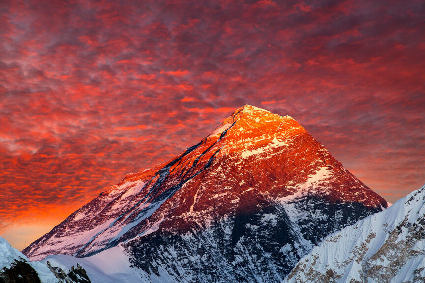A snow-capped mountain peak is illuminated by a vibrant red and orange sunset, with a sky full of scattered clouds—a perfect backdrop for an unforgettable REI Adventure.
