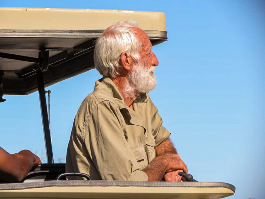 An older man with white hair and beard, wearing a tan shirt, stands under a shade canopy looking into the distance against the clear blue sky, likely reflecting on his time spent in Namibia working on wildlife conservation efforts.