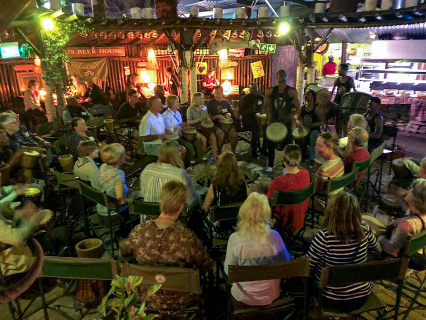 A group of people, including wildlife conservationists, sit in a circle, playing drums and musical instruments at an indoor gathering. The setting appears to be a casual, rustic venue with dim lighting. It's a special event inspired by Namibia's rich cultural heritage.