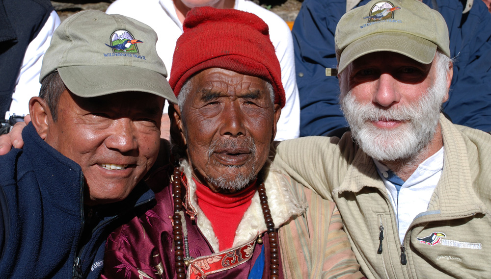 Three men are seated closely together, all smiling. The man in the middle wears a red hat and traditional clothing, while the men on each side wear outdoor gear and caps with a logo. This joyful snapshot would be perfect for any family photobook, capturing their bond and shared adventure in Nepal.