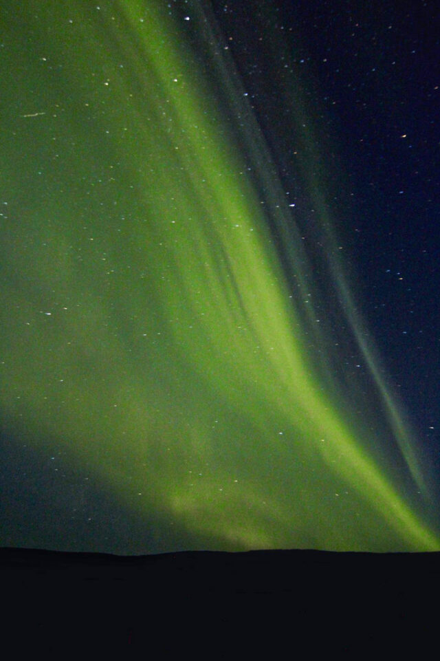 Green aurora borealis streaking across a dark, starry night sky, with a faint outline of the horizon at the bottom. Such breathtaking displays are often seen during hikes in Iceland, where the Northern Lights dance vividly overhead.