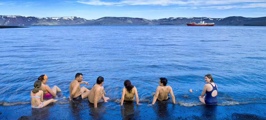Seven people in swimwear sit on a black sand shore, facing a calm blue body of water with a distant red and white ship. Snow-capped mountains are visible on the horizon under a clear sky, as if they've found solace in one of nature's heavenly hot springs.