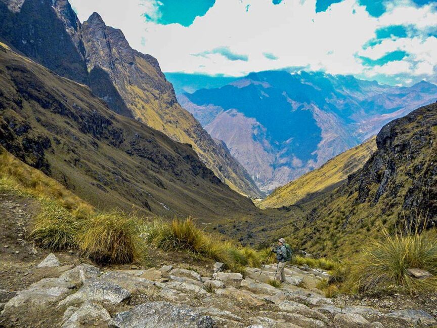 A hiker walks along a rocky path surrounded by steep mountains and valleys under a blue sky with scattered clouds, similar to the breathtaking vistas of the Inca Trail.