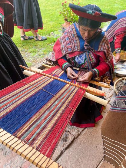 A person in traditional clothing weaves a colorful textile on a loom, with a focus on red and blue threads. In the Sacred Valley of Peru, other similarly dressed individuals are visible in the background, discovering ancient techniques.