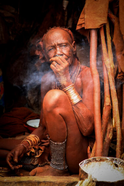 A person sits on the ground surrounded by wooden poles, wearing traditional attire and jewelry, and appears to be deep in thought while exhaling smoke, the scene imbued with the rich colors of the wild.