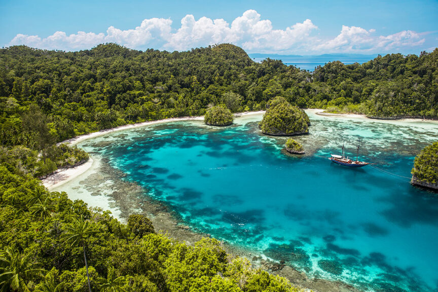 Aerial view of a tropical bay with clear turquoise water, lush green trees, small islands, and a boat anchored near the shore—an idyllic scene perfect for trips to book with advance booking for the best experience.