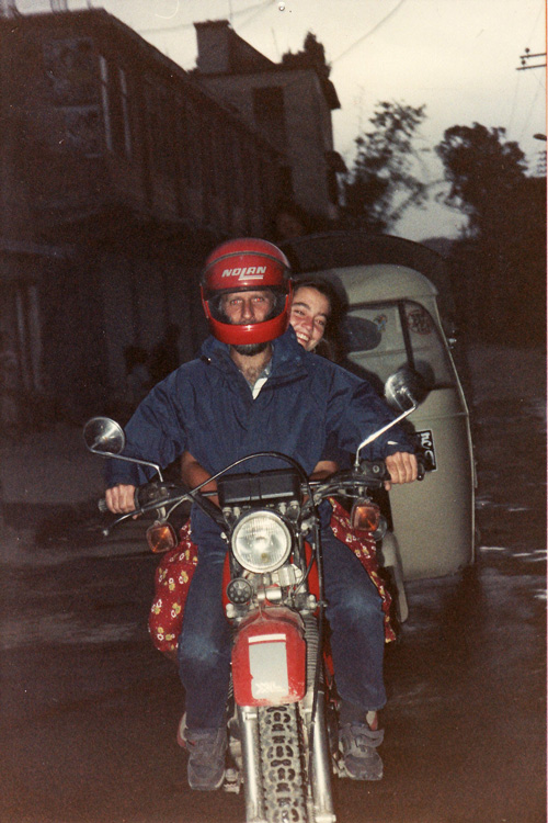 Two individuals ride a motorcycle with the driver wearing a red helmet and the passenger smiling, seen on a street in Nepal with buildings and a vehicle in the background—an ideal moment for any family photobook.