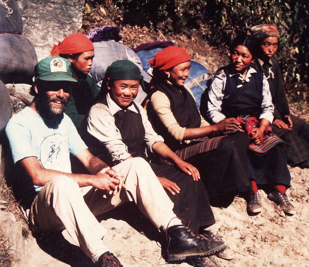 A group of six people, consisting of five women and one man, are sitting outdoors on the ground in Nepal. They are dressed in traditional and casual clothing, appearing to be enjoying a moment together that could easily belong in a cherished family photobook.