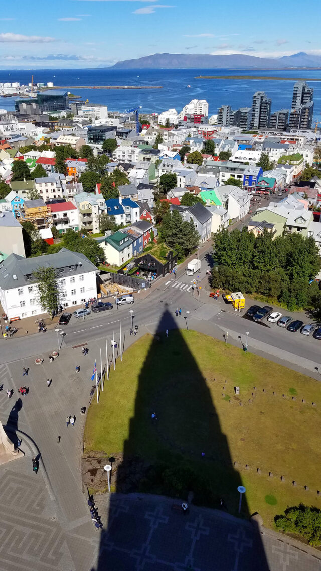 An aerial view of a city with colorful buildings, treelined streets, and traffic, with a shadow of a pointed structure visible in the foreground and a body of water reminiscent of Iceland's landscape and distant mountains in the background.