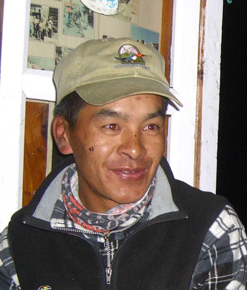 A person wearing a green cap and a checkered scarf smiles while sitting indoors in front of a window, with photographs on the wall behind them, capturing moments from their family photobook.