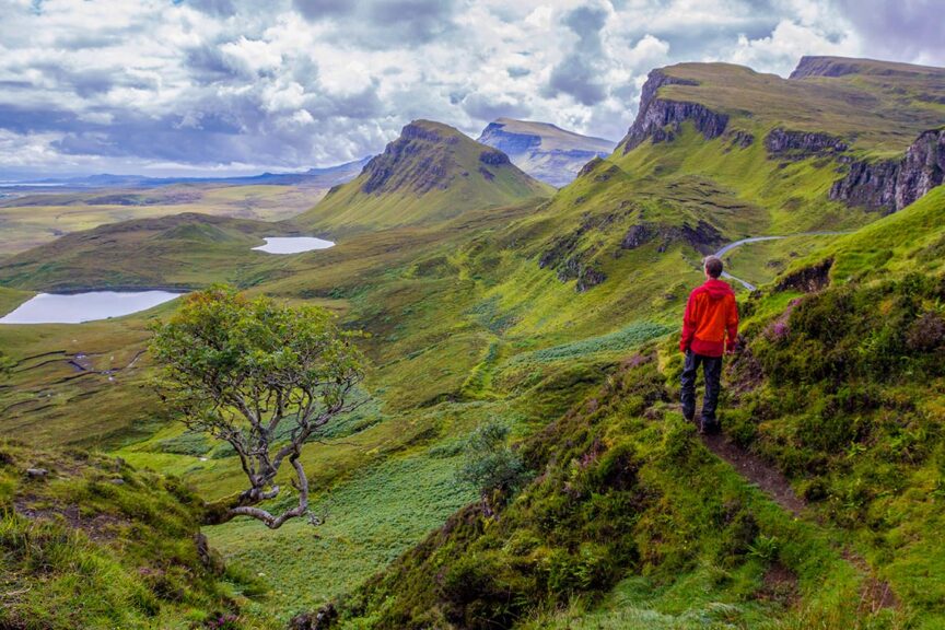 A person in a red jacket stands on a trail overlooking a lush green valley with hills, cliffs, and two small lakes under a cloudy sky, as canceled trips have led to an appreciation for local adventures.