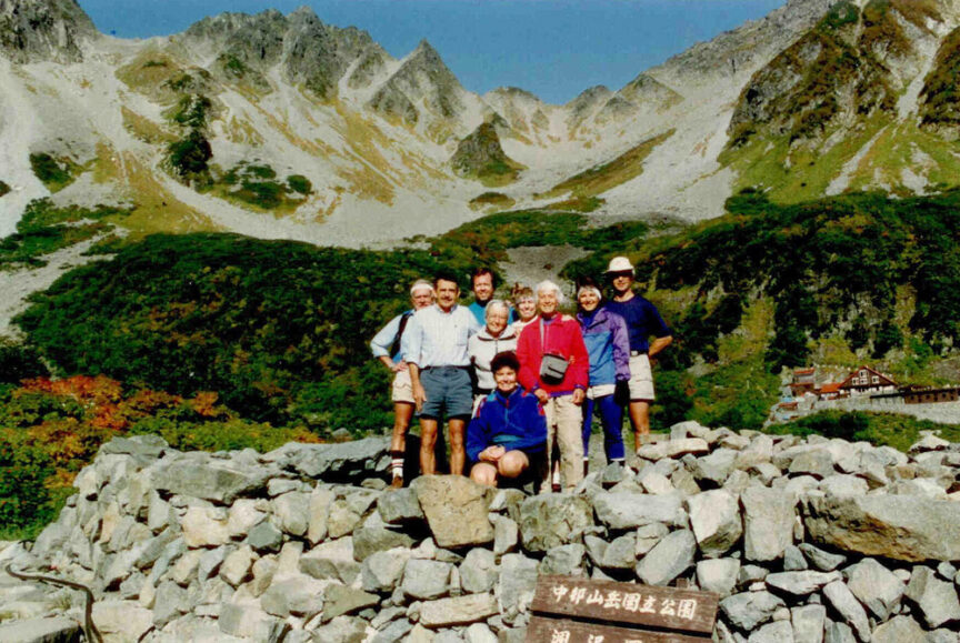 A group of eight people in outdoor attire pose for a photo on a stone structure with a mountainous backdrop, capturing their journey. A sign in the foreground has text in Japanese, reminiscent of an ancient haiku.