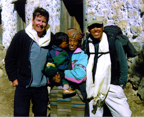 Four people posing outside in front of a stone building, set against the serene backdrop typical to Nepal. One person holds a child, and two others wear white scarves around their necks. The individuals are dressed warmly and appear to be in a mountainous area, perfect for a family photobook.