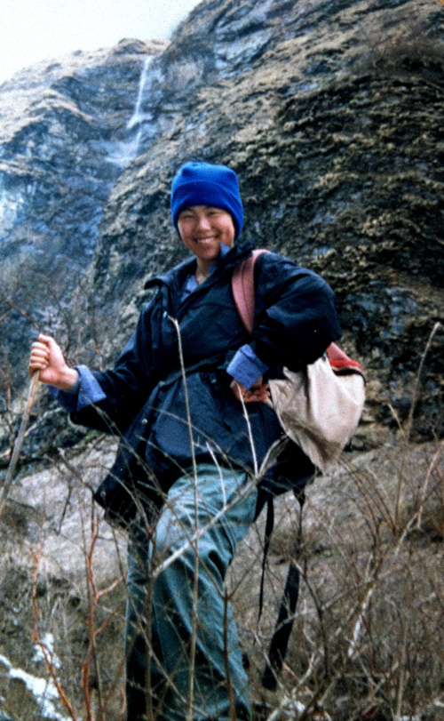 Person wearing outdoor gear and blue hat stands on a rocky, grassy hillside in Nepal with a trekking pole and a backpack, smiling at the camera, perfect for the family photobook.