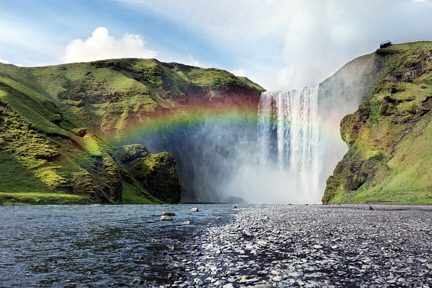 A large waterfall cascades down a cliff, surrounded by green hills with a rainbow visible in the mist at the base. Rocky terrain and a shallow river are in the foreground under a partly cloudy sky, making it a perfect spot to visit for those who love waterfalls and travel.