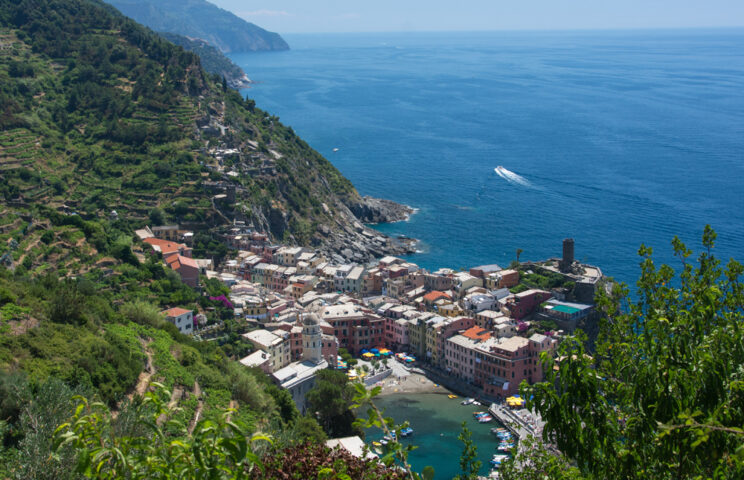 A small coastal village is nestled between the green hills of the Apennines and the blue sea, with boats and buildings visible along the shoreline, inviting a sense of adventure.
