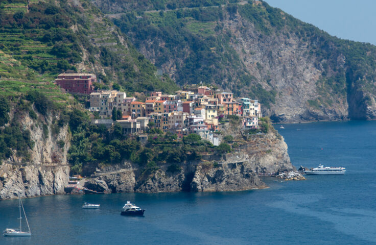 A coastal village with colorful buildings perched on a cliff, surrounded by the rugged terrain of the Apennines, overlooks a calm blue sea dotted with yachts and sailboats, promising an adventure at every turn.