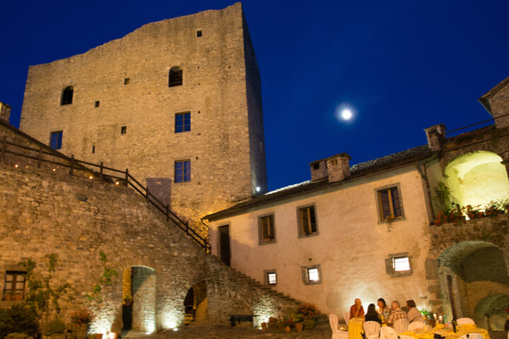 A group of people enjoy a dinner at an outdoor courtyard near an ancient stone building under a clear night sky with a visible moon, ready to share tales of their adventures in the Apennines.