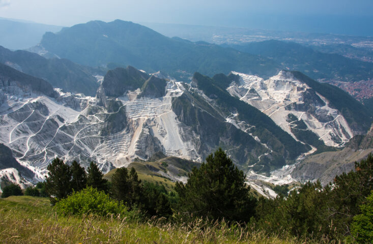 Aerial view of marble quarries in the Apuan Alps, Italy, with terraces carved into the mountains and greenery in the foreground, blending seamlessly into the hazy Apennines in the distance—an adventure waiting to be explored.