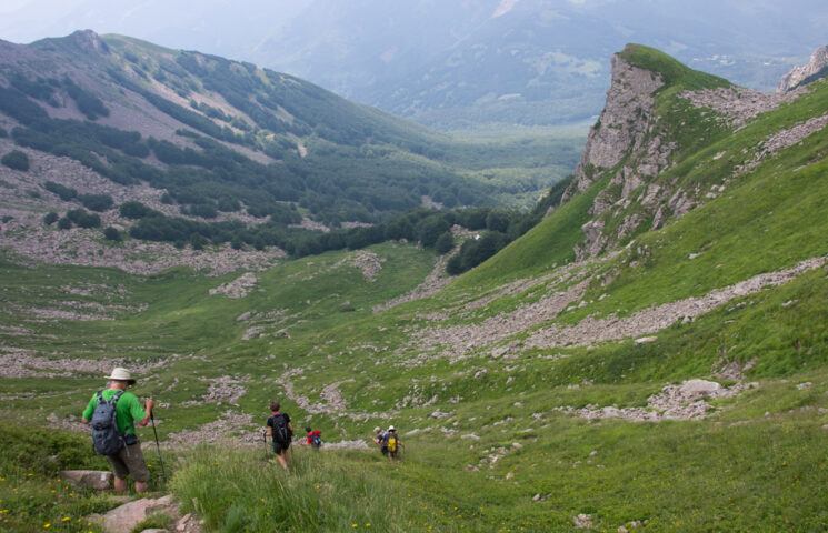 Hikers embark on an adventure down a green mountain trail in the Apennines, surrounded by rocky and grassy slopes, with a vast valley and distant mountains in the background.