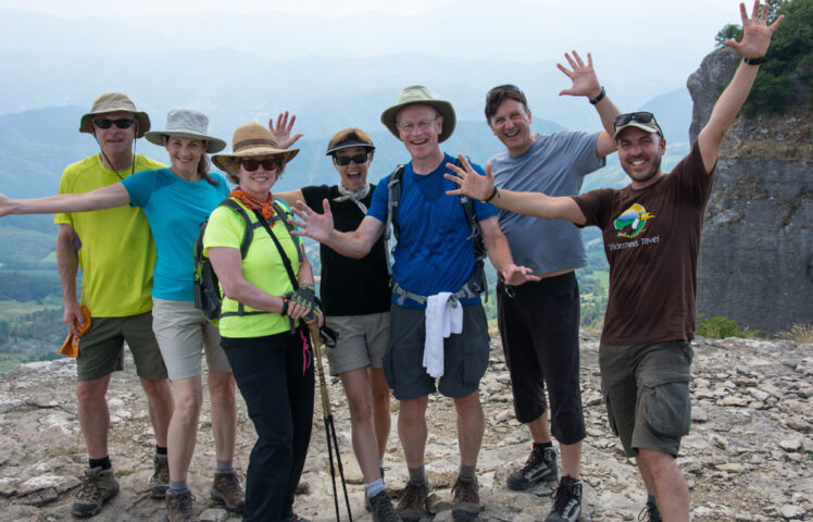 A group of seven people, wearing hiking gear and hats, stand on a rocky ledge with their arms raised, smiling at the camera. A scenic Apennines mountain landscape is visible in the background, capturing their adventure perfectly.