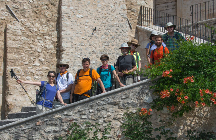 A group of nine hikers, wearing casual outdoor clothing and hats, stand on stone steps near a rustic building adorned with a flowering vine. One person holds a camera on a selfie stick, capturing the spirit of their Apennines adventure.