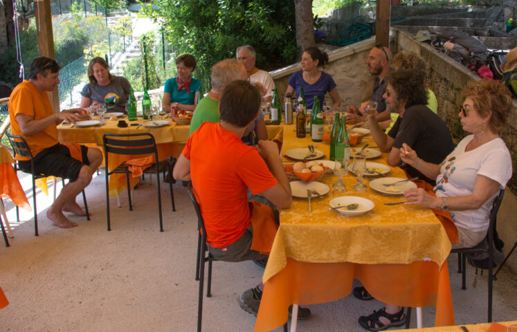 A group of people are seated around tables outdoors, enjoying a meal together after an adventure in the Apennines. The tables, covered with yellow tablecloths, are adorned with various bottles and plates. Trees and a garden create a serene backdrop.