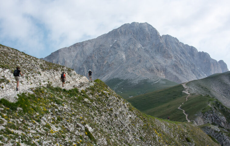 Hikers on a mountainous trail with a rocky peak in the background, under a partly cloudy sky, embark on their adventure through the enchanting Apennines.