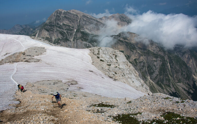 Hikers embark on an adventure, traversing a rocky, snow-covered mountain trail with the cloud-covered peaks of the Apennines in the background on a clear day.